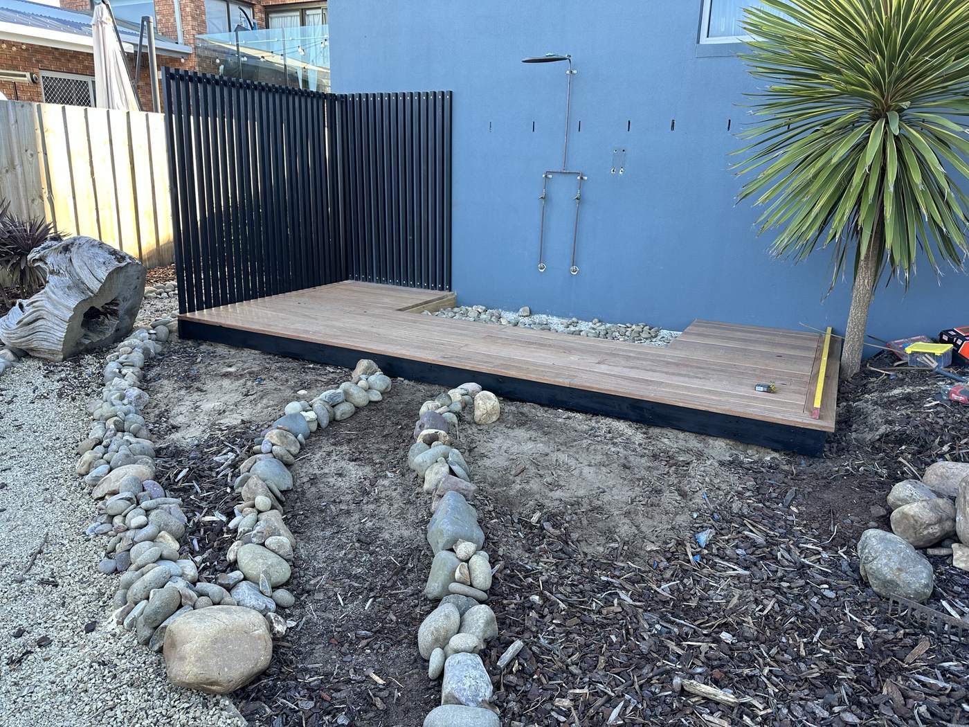 L-shaped hardwood deck around a river-stone shower base, with a black batten privacy screen behind